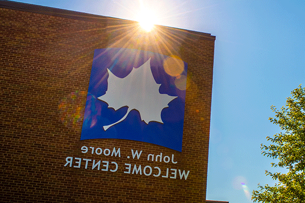 A brick building on the right of the image with a large Indiana State logo and “Welcome Center” text with the sun peeking over the building. A tree is visible in the left side of the frame with a blue sky.